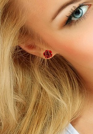 Red rose-shaped stud earring on a model's ear, featuring intricate detailing and a glossy finish. Curled blonde hair partially visible.