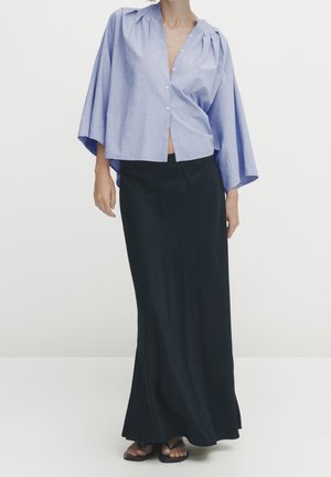 Kobieta ubrana w luźną, jasnoniebieską, zapinaną na guziki bluzkę z szerokimi rękawami oraz długą czarną spódnicę, zestawioną z czarnymi sandałami.