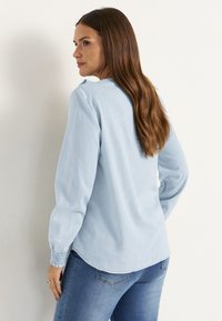 Lichtblauwe blouse met lange mouwen van zachte stof, met smock manchetten en een ronde zoom. G gedragen met blauwe jeans, wat een casual stijl laat zien.