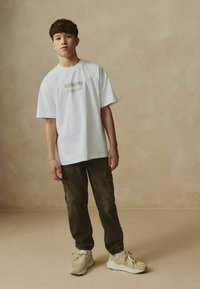 T-shirt bianca a maniche corte con la scritta "ISCRIVITI" stampata in oro, abbinata a pantaloni cargo larghi scuri e sneakers beige. Tessuto morbido e fit rilassato.