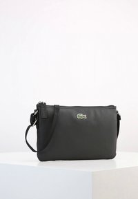 Svart nylonväska att bära över kroppen med strukturerad yta, försedd med dragkedja och en liten grön krokodil-logotyp. Justerbar axelrem.