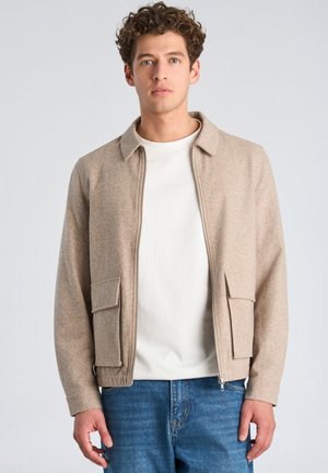 Junger Mann mit lockigem Haar, der eine beige Reißverschlussjacke über einem weißen Hemd und blauen Jeans trägt und vor einem schlichten hellen Hintergrund steht.