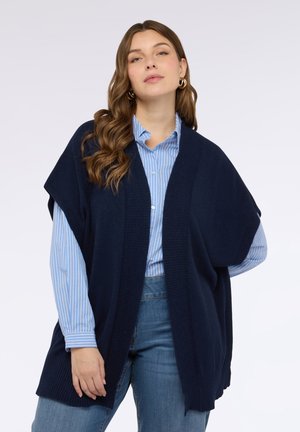 Gilet lavorato a maglia blu navy con davanti aperto, bordi a coste e vestibilità ampia, indossato sopra una camicia a righe azzurre con bottoni e jeans di denim.