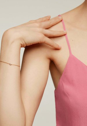 Vestido de satén rosa con tirantes finos, adornado con una delicada pulsera de oro que lleva una pequeña gema central, resaltando la textura suave de la piel.