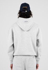 Felpa grigia con cappuccio, caratterizzata da una vestibilità rilassata e polsini a costine. Un berretto blu navy con testo ricamato completa l'outfit.