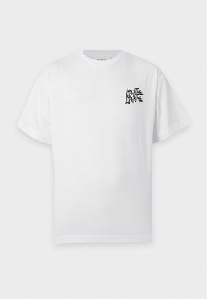 Homme debout portant un t-shirt blanc avec un petit texte noir sur la poitrine, un pantalon large sombre et des chaussures claires sur un fond uni.