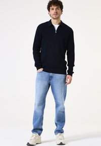 Navy gebreide trui met een ritskraag, gecombineerd met lichtblauwe denim jeans en crèmekleurige sportschoenen. Heeft een ribbeltextuur en een relaxte pasvorm.
