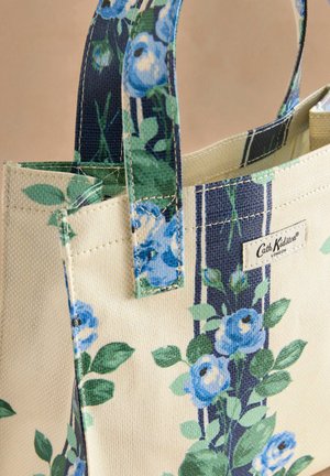 Torba za plažu s uzorkom u krem boji s plavim cvjetnim dizajnom i zelenim listovima, s okomitim tamnoplavim prugama i brendiranim etiketom. Tekstuirana tkanina.