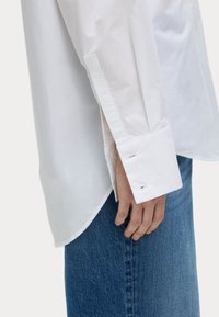 Weißes, langärmliges Hemd aus glattem Baumwollstoff mit seitlichem Schlitz und geknöpften Manschetten, kombiniert mit blauen Jeans.