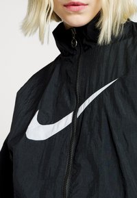 Črna Nike jakna z visokim ovratnikom, zadrgo in izstopajočim belim logotipom swoosh spredaj. Teksturni, lahki material.