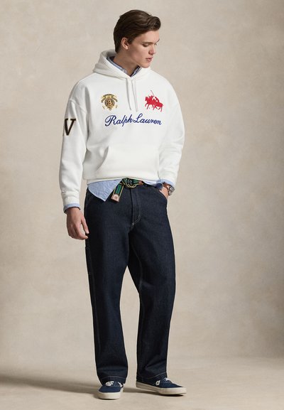 Młody mężczyzna w białej bluzie z kapturem Ralph Lauren, niebieskiej koszuli, ciemnych dżinsach i trampkach, stojący z jedną ręką w kieszeni na jednolitym tle.