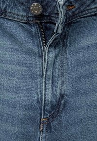 Gros plan sur un jean en denim bleu avec une fermeture éclair défaite et un bouton métallique à la ceinture.