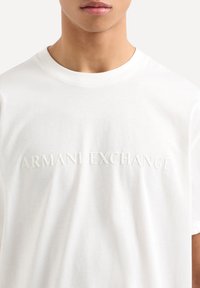 T-shirt bianca in cotone con logo in rilievo "ARMANI EXCHANGE" sul petto. Maniche corte e scollatura rotonda. Texture liscia.