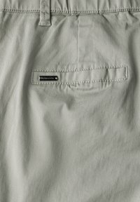 Pantalons vert pâle avec une poche arrière ornée d'un logo argenté, texture lisse et légère brillance. Design simple avec des accents cousus.