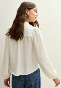 Femme aux longs cheveux bruns portant un chemisier blanc à manches longues et un jean bleu, de profil devant un fond clair.