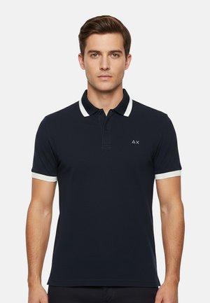 Uomo con una polo nera con rifiniture bianche su colletto e maniche, in piedi davanti a uno sfondo chiaro e uniforme.