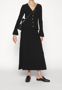 Černá pletená souprava se žebrovanou strukturou, která zahrnuje cardigan s V-krkem a zlatými knoflíky a odpovídající dlouhou sukni. Doplněno černými botami na slip-on.