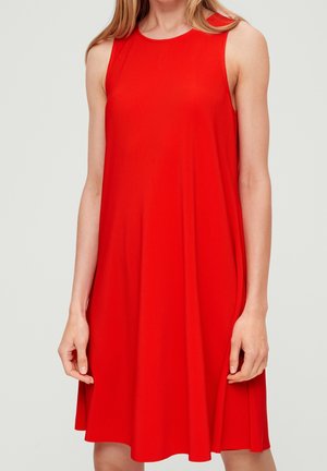 Nainen pukeutuneena hihattomaan kirkkaan punaiseen ribbineulottuun A-linjamekkoon, seisomassa käsien rentona sivuilla vaaleaa taustaa vasten.