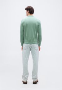 Homme debout de dos portant une chemise à manches longues vert clair, un jean bleu clair et des chaussures marron sur un fond blanc uni.