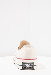 Plátěné tenisky v off-white s červeným pruhem a černou patou s bílým textem. Plochá podrážka a zaoblený design špičky.