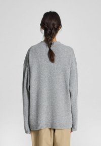 Šedý oversize svetr s ribbed lemováním a jemnou strukturou. Má spadlá ramena a uvolněný střih, nošený přes béžové kalhoty.