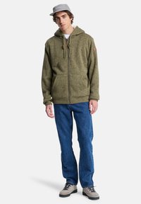Groene hoodie met vlekken, met rits en een trekkoordkap, voorzakken en een logo op de linkermouw, gecombineerd met blauwe jeans en een grijze pet.
