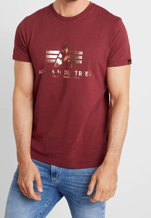 Mężczyzna w bordowej koszulce Alpha Industries z metalicznym logo i niebieskich dżinsach, stojący na jasnym, jednolitym tle.