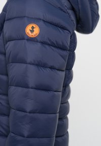 Marineblauwe pufferjack met quilted segmenten en een oranje cirkelvormig logo. Glad, glanzend materiaal met een capuchon.
