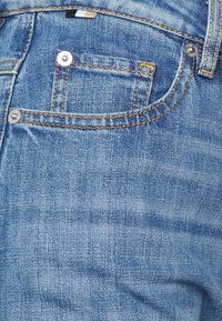 Jeans i ljusblå denim med en något urtvättad textur. Har en liten framficka och metallknappar med oranga sömnadsdetaljer.