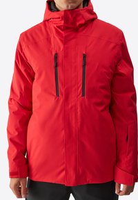 Giacca isolante rossa con colletto alto, cappuccio regolabile, due tasche sul petto con zip e polsini in velcro. Esterno liscio e impermeabile.