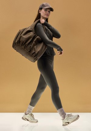 Frau in dunkler Sportkleidung und Mütze, die mit einer großen braunen Tasche über der Schulter vor einem beigen Hintergrund geht.