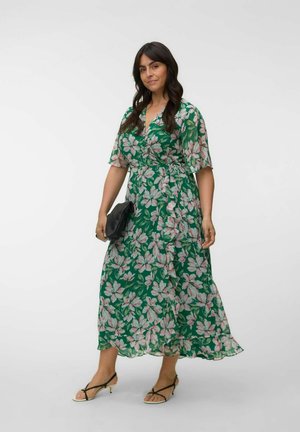 Kvinde i grøn blomsterprintet omslagskjole, der holder en sort clutch og iført sorte sandaler med lav hæl og stropper, stående mod en ensfarvet baggrund.