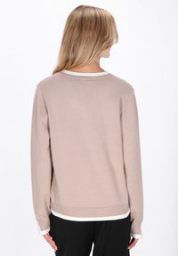 Femeie cu păr lung și blond, purtând un pulover bej cu margini albe și pantaloni negri, văzută din spate pe un fundal alb simplu.