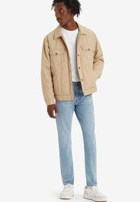 Quiltet beige jakke med to frontlommer, trykknapper og krage, båret over en hvit t-skjorte, paret med lyseblå jeans og hvite joggesko.