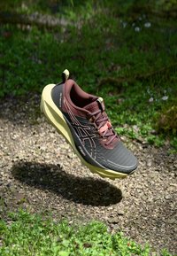 Brown and black lightweight running shoe with a textured upper, yellow sole, and reinforced toe, shown in mid-air above a gravel path.
