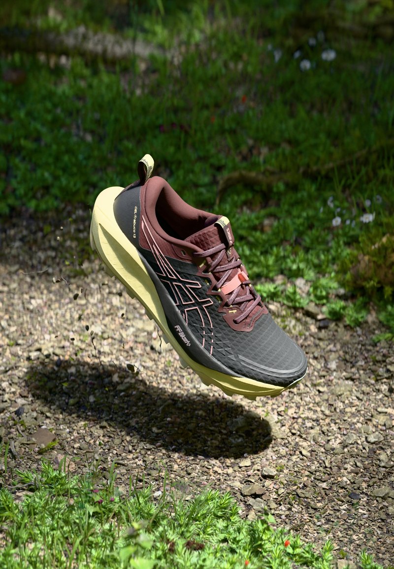 Brown and black lightweight running shoe with a textured upper, yellow sole, and reinforced toe, shown in mid-air above a gravel path.