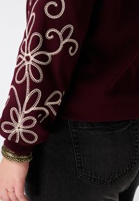 Bordeaux sweatshirt met een gestructureerd gouden bloemenborduurpatroon op de mouw, gecombineerd met donkere spijkerbroeken.