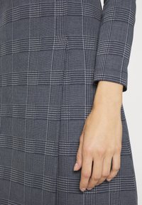 Granatowa sukienka w kratę, wykonana z teksturowanej tkaniny z małymi kwadratami i wzorem houndstooth; rękawy trzy czwarte i dopasowany krój.