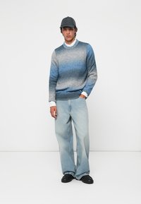 Jeune personne portant un pull dégradé bleu-gris, un jean large clair, des chaussures noires, une chemise blanche et une casquette noire, se tenant devant un fond blanc.