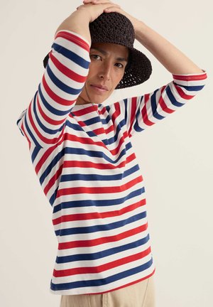 Persoană purtând o cămașă cu dungi roșii, albe și albastre și o pălărie neagră împletită, ridicând brațele pentru a-și ajusta pălăria pe un fundal simplu.