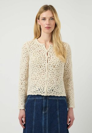 Donna che indossa un cardigan floreale all'uncinetto color crema abbinato a una gonna di denim blu, in piedi davanti a uno sfondo chiaro e semplice.