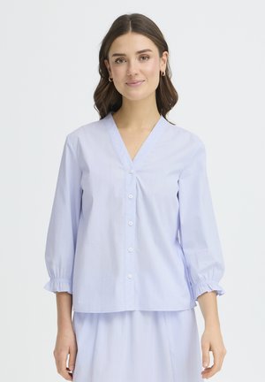 Mladá žena s hnedými vlasmi po plecia, oblečená v svetlomodrej blúzke na gombíky s tromištvrťovými volánovými rukávmi a zodpovedajúcej sukni.