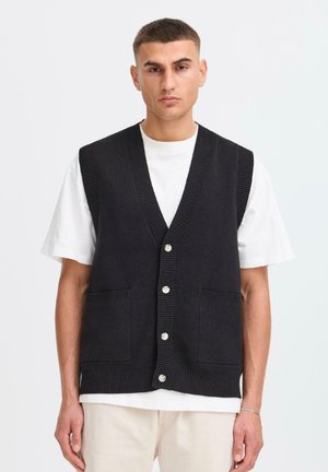 Een man in een zwarte gebuttonde gebreide vest over een wit shirt met korte mouwen en lichte broek, staande tegen een effen lichte achtergrond.