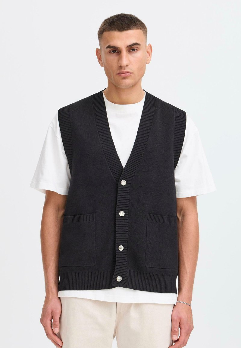 Een man in een zwarte gebuttonde gebreide vest over een wit shirt met korte mouwen en lichte broek, staande tegen een effen lichte achtergrond.