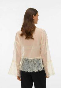 Proua pruunide pikkade juustega naine, kes kannab läbipaistvast beežist pluusi pitsist ääristusega varrukatel ja servadel, mustade pükstega, seljaga vaataja poole.