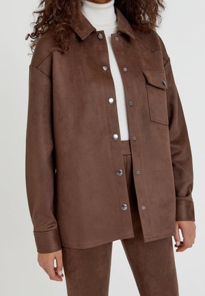 Kvinde iført brun ruskindsknapjakke og matchende bukser over en hvid ribbet rullekravesweater.