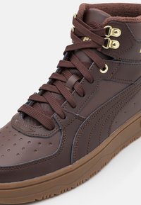 Brązowe trampki typu high-top wykonane ze skóry, z perforowanym noskiem, teksturowanymi akcentami, złotymi oczkami i brązowymi sznurówkami na gumowej podeszwie.