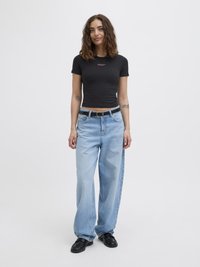 Schwarzes, gekürztes T-Shirt mit pinkem Text, hellblaue weit geschnittene Jeans und schwarze Schuhe. Die Haare sind wellig und schulterlang. Ein schlichter Hintergrund.