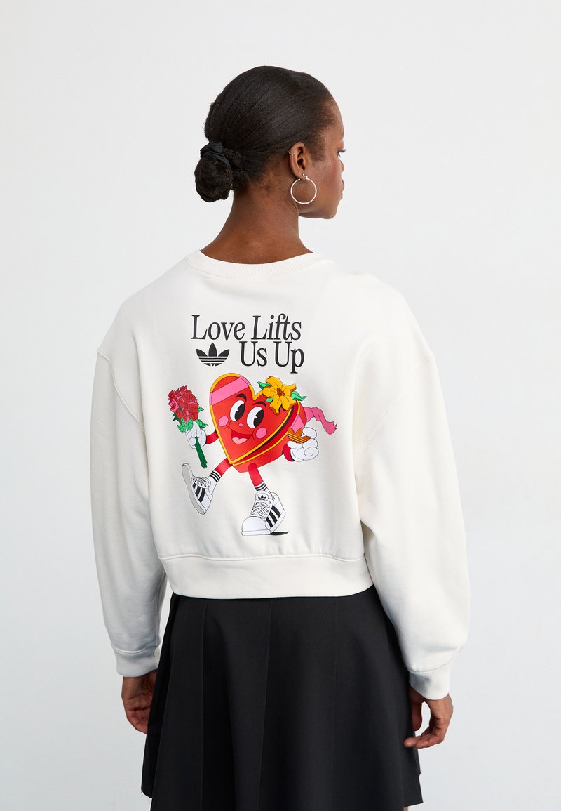 Λευκό κοντό φούτερ με σχέδιο καρδιάς ήρωα στην πλάτη, κείμενο "Η Αγάπη Μας Υψώνει" και μαύρες λεπτομέρειες. Λείο ύφασμα και άνετη εφαρμογή.