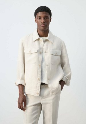 Mann trägt eine cremefarbene Cordjacke und passende Hose, steht mit einer Hand in der Tasche vor einem schlichten hellen Hintergrund.